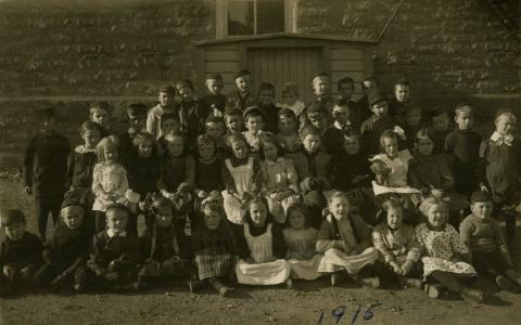 Black and white photograph of school children behind Victoria School. The class portrait has four rows, both boys and girls. The first row is seated. Behind them is a wooden shed. At the bottom is "1915" written in blue ink.